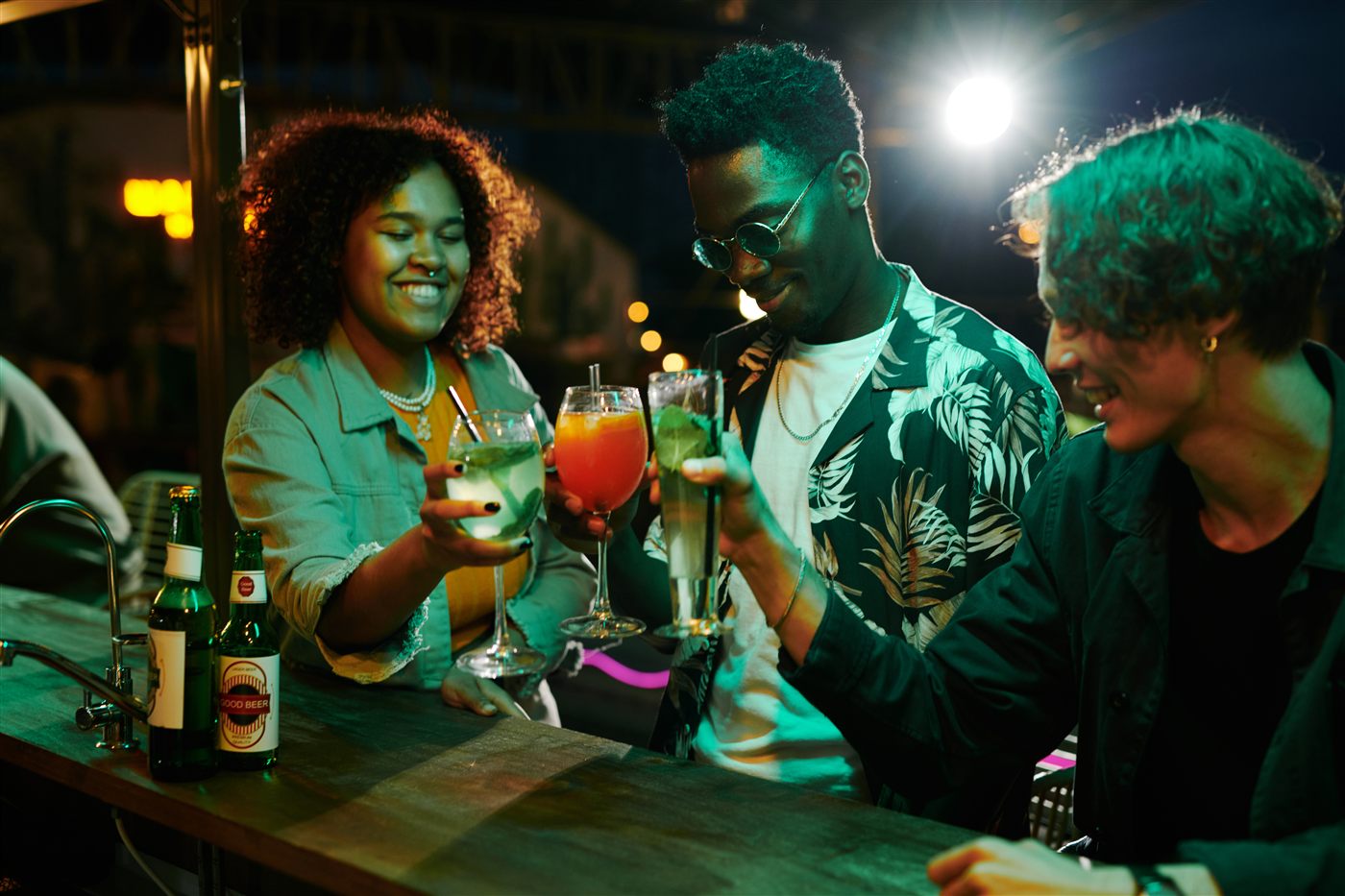 Three young friends sharing drinks together at night, reflecting the social mix of locals, diaspora, and visitors moving through the same space.
